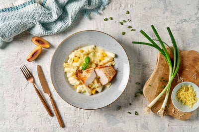 Ein Teller Pasta mit Hähnchenscheiben, dazu Pfirsiche und Frühlingszwiebeln, elegant mit Besteck angerichtet auf einer strukturierten Unterlage – dieses erlesene Gericht ließe sich problemlos in ein farbenfrohes „Schlemmen ohne Grenzen“ von Hofmanns Shop packen und ist die ideale Mahlzeit für unterwegs.