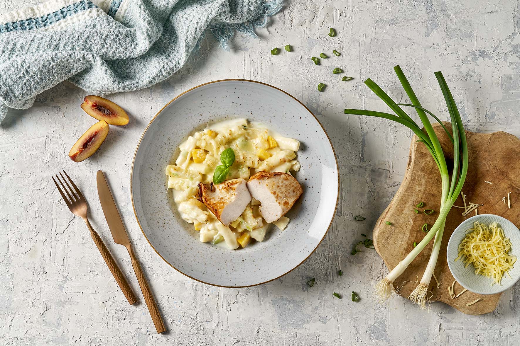 Ein Teller Pasta mit Hähnchenscheiben, dazu Pfirsiche und Frühlingszwiebeln, elegant mit Besteck angerichtet auf einer strukturierten Unterlage – dieses erlesene Gericht ließe sich problemlos in ein farbenfrohes „Schlemmen ohne Grenzen“ von Hofmanns Shop packen und ist die ideale Mahlzeit für unterwegs.