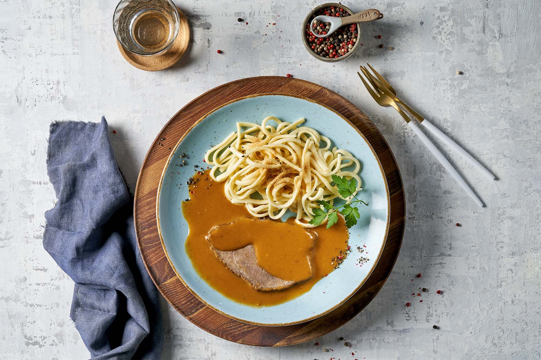 Ein Gericht aus Hofmanns Shop, bekannt als Omas Verwöhnmenü, besteht aus Nudeln mit pikanter Soße und Fleisch, garniert mit Kräutern, um die Essenz traditioneller Lieblingsgerichte einzufangen. Dazu gibt es ein Getränk, eine Serviette und eine Pfefferschale.