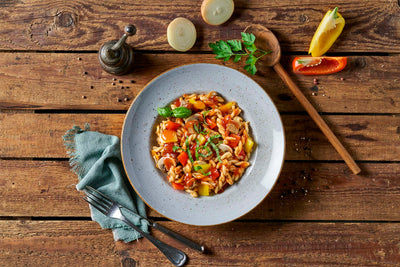 Eine Schüssel Pasta mit Gemüse auf einem rustikalen Holztisch, begleitet von Besteck und Gewürzen, spiegelt die Qualität der HOFMANNs Überraschungsbox wider.