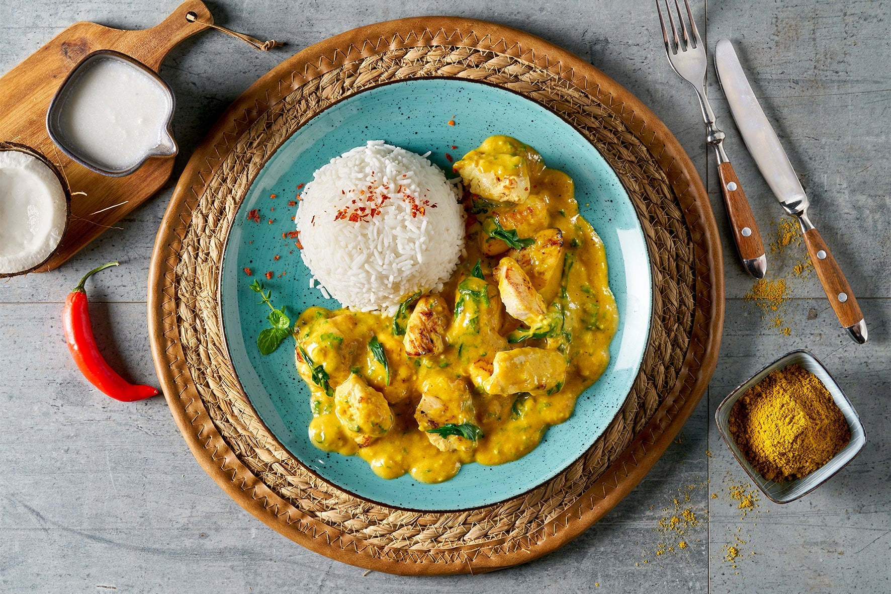 Ein blauer Teller mit weißem Reis und cremigem Curry-Hühnchen, garniert mit Kräutern, entführt Sie auf eine Große asiatische Genussreise, mit freundlicher Genehmigung von Hofmanns Shop.