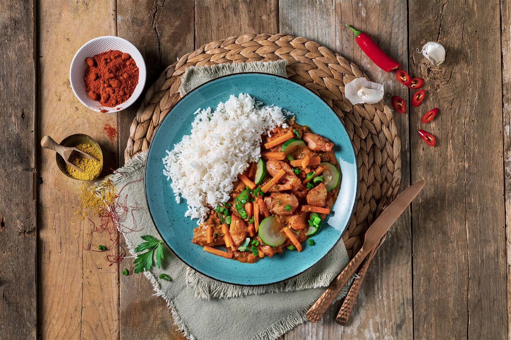 Teller „Große asiatische Genussreise“ von Hofmanns Shop, mit Reis mit Curry, Gemüse und Hühnchen auf einem rustikalen Holztisch, umgeben von Gewürzen und Chili – eine exotische kulinarische Reise, die die Sinne kitzelt.