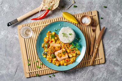 Hofmanns Shop Fisch-Box: buntes Currygericht mit Reis auf blauem Teller, serviert auf einer Bambusmatte.