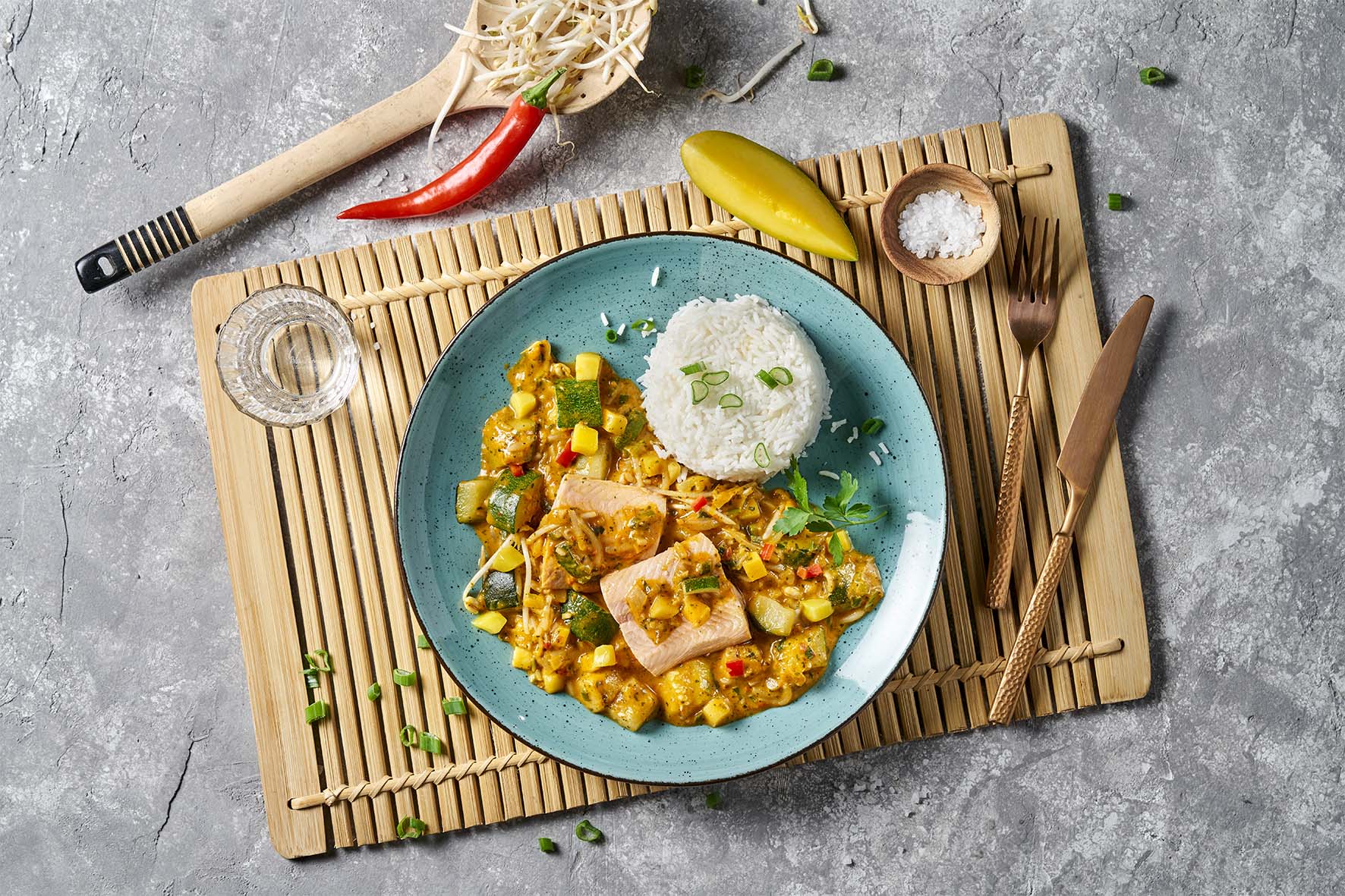 Hofmanns Shop Fisch-Box: buntes Currygericht mit Reis auf blauem Teller, serviert auf einer Bambusmatte.