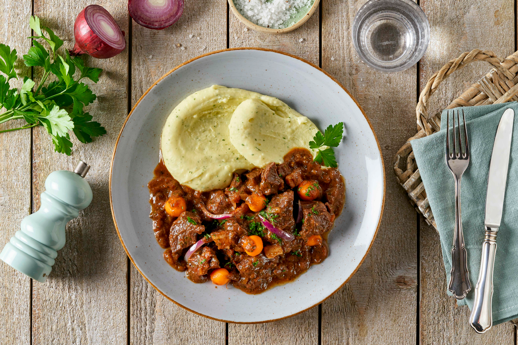 Ein Teller Boeuf à la Bourguignon von Hofmanns Shop, serviert mit Kartoffelstampf und Karotten, garniert mit Petersilie, auf einem Holztisch platziert.