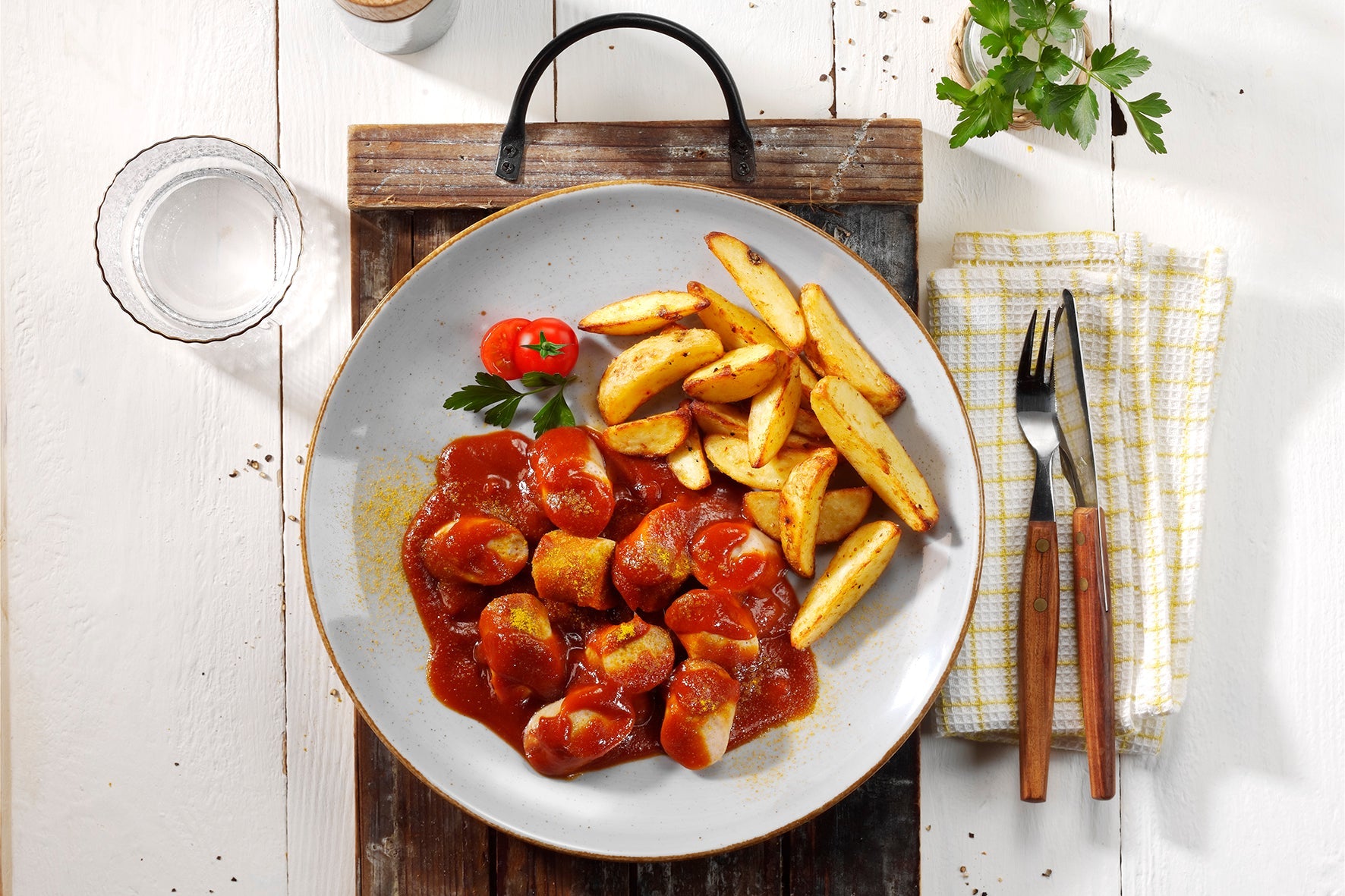 Eine HOFMANNs Neuheiten Currywurstplatte mit Pommes, Petersilie, Kirschtomate, Utensilien und Wasser von Hofmanns Shop.