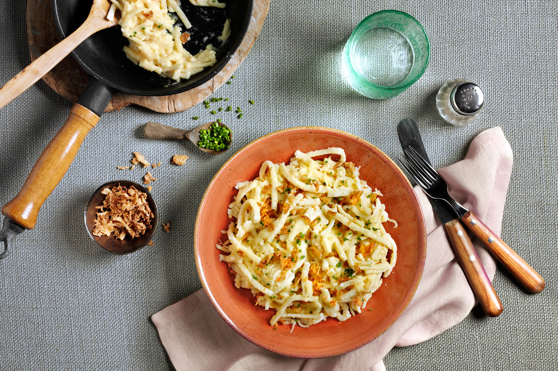 Teller mit Käse-Pasta mit Schnittlauch, Röstzwiebeln, Besteck, einem Glas und einer Pfanne auf grau strukturiertem Untergrund – ein wahrer HOFMANNs Osterbox-Genuss aus Hofmanns Shop, der Restaurantqualität in Ihre Festtagsgerichte bringt.