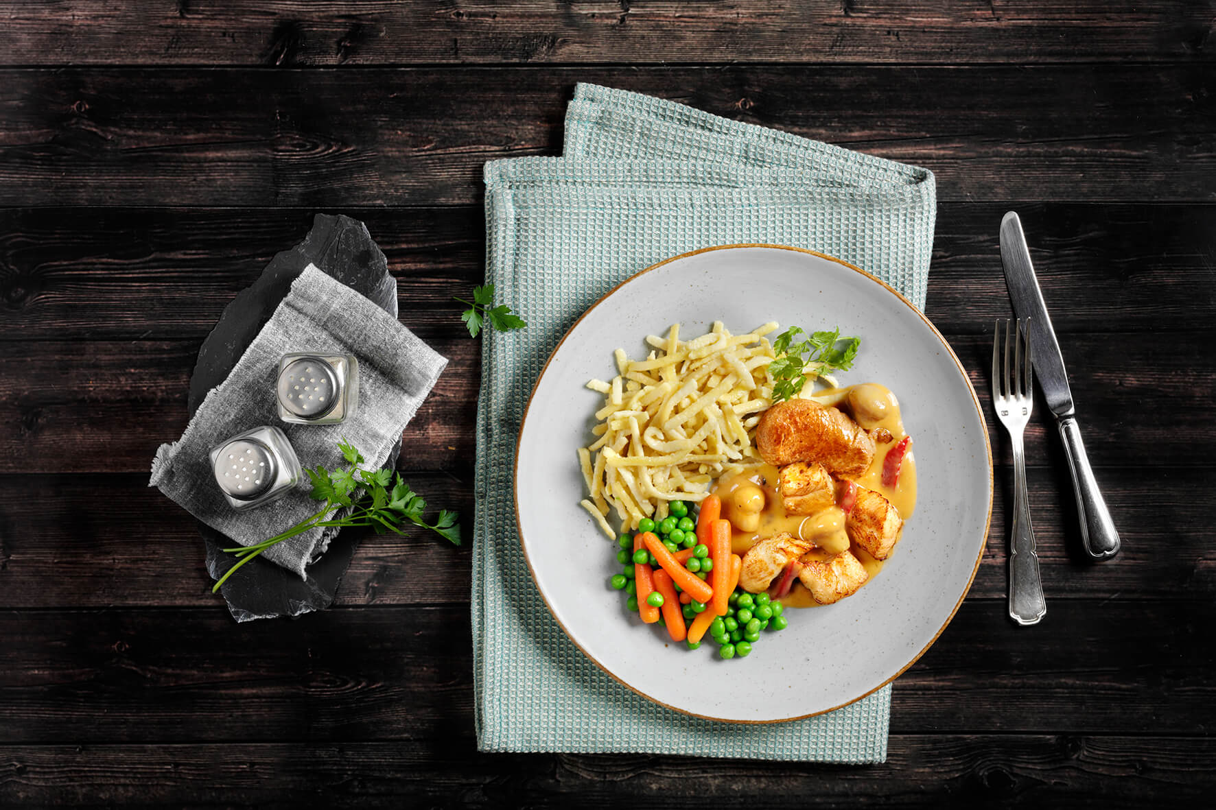 Die HOFMANNs Osterbox von Hofmanns Shop besteht aus Hähnchen mit Nudeln, Erbsen und Karotten auf einer grünen Unterlage und strahlt Restaurantqualität aus. Besteck und Gewürzstreuer daneben runden das Festtagsgericht-Ambiente ab.