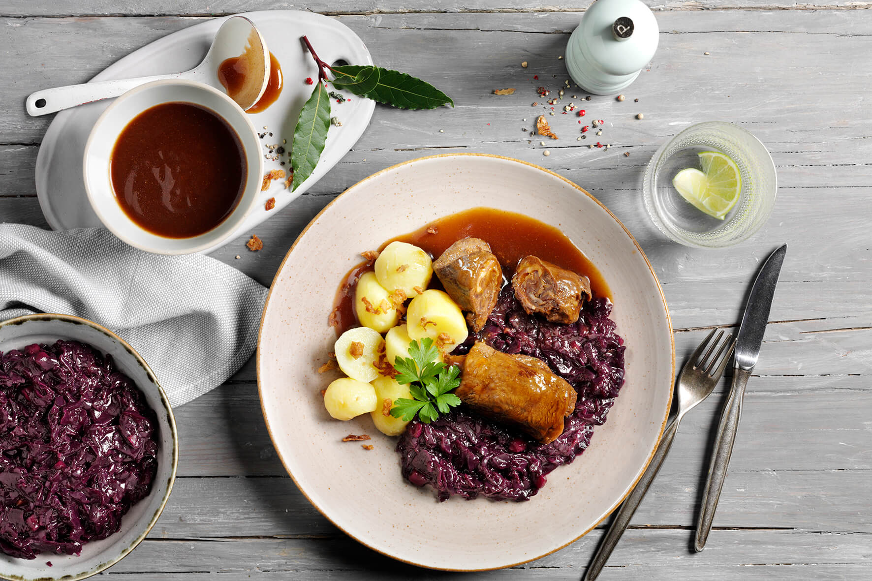 Ein Teller mit „Unsere Küchenhighlights“ von Hofmanns Shop, bestehend aus Rinderrouladen, Rotkohl und Kartoffeln mit Bratensoße, wird auf einem Holztisch präsentiert, daneben steht ein Glas Zitronenwasser.